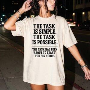 May include: A beige short-sleeved t-shirt featuring black text that reads: THE TASK IS SIMPLE. THE TASK IS POSSIBLE. THE TASK HAS BEEN "ABOUT TO START" FOR SIX HOURS. The shirt has a crew neckline and a relaxed, loose-fitting silhouette.
