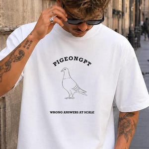 May include: A white short-sleeved t-shirt featuring a black line drawing of a pigeon in the center. Above the pigeon, the text reads PIGEONGPT in a curved font, and below the bird, the text reads WRONG ANSWERS AT SCALE in a bold font.