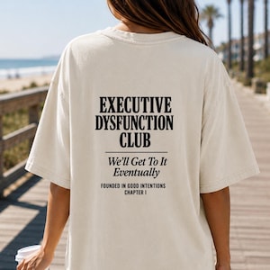 May include: A cream-coloured t-shirt featuring the black text "EXECUTIVE DYSFUNCTION CLUB", the phrases "We'll Get To It Eventually" and "FOUNDED IN GOOD INTENTIONS CHAPTER I". The shirt has a relaxed, oversized style.