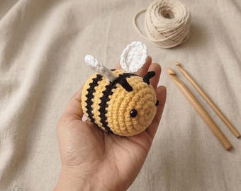 May include: A handmade crochet bee with yellow and black stripes, white wings, and black eyes. The bee is held in a hand, with a ball of yarn and two wooden crochet hooks in the background. The bee is a cute, handcrafted item.