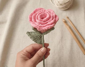 May include: A handmade crochet rose in shades of pink, green, and gray. The rose is held in a hand, with a ball of yarn and crochet hooks in the background. The rose has a detailed petal design and a green stem and leaves. The logo "Soft Petal Crochet" is in the bottom left corner.