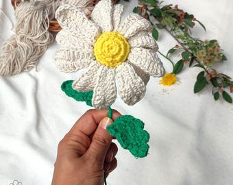 May include: A handcrafted crochet daisy flower featuring white petals and a bright yellow center. The flower is attached to a thin green stem with two textured green leaves. The design includes a small logo in the corner reading Soft Peta Crochet.