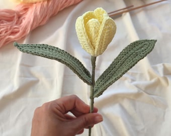 May include: A handcrafted crochet tulip flower featuring a pale yellow bloom and two long, pointed sage green leaves attached to a thin, wrapped stem. The texture of the yarn is visible throughout the petals and the foliage of the flower.