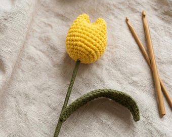May include: A handmade crochet tulip in bright yellow, with a green stem and leaf. Two wooden crochet hooks and a ball of yarn are in the background. The flower is set against a neutral linen background.