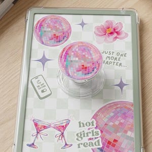 May include: A clear phone case with a green border, featuring a pop socket and stickers. The stickers include disco balls, a pink flower, martini glasses, and the text "hot girls read" and "just one more chapter".