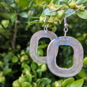 May include: A pair of silver-coloured, open-square earrings with a glittery, translucent appearance. The earrings have a hook closure and are embellished with blue and gold flecks. The earrings are hanging in front of a green leafy background.