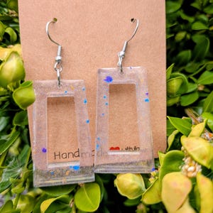 May include: Clear rectangular earrings with silver hooks, filled with glitter and colourful specks. The earrings are attached to a brown card with the words "Handmade with love" printed on them.