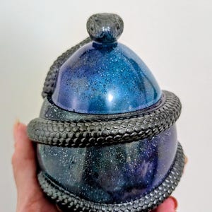 Könnte beinhalten: Ein kugelförmiges Gefäß mit Deckel in einer tiefblauen und schwarzen, galaxieartigen Optik mit silbernen Glitzerakzenten. Eine texturierte, dunkelgraue, skulptierte Schlange windet sich um den Körper des Gefäßes, wobei ihr Kopf als Griff auf dem Deckel ruht.