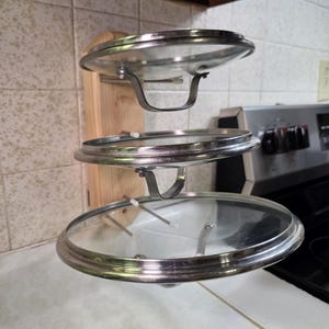 May include: A wall-mounted wooden rack featuring three horizontal metal rods that support three circular glass pot lids with stainless steel rims and metal handles. The rack is constructed from light-colored wood and is mounted vertically.