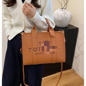 May include: A brown tote bag with top handles and a shoulder strap. The bag has the words "TOT" and "MARC" printed on the front. The bag is made of a smooth material and has gold-colored hardware.