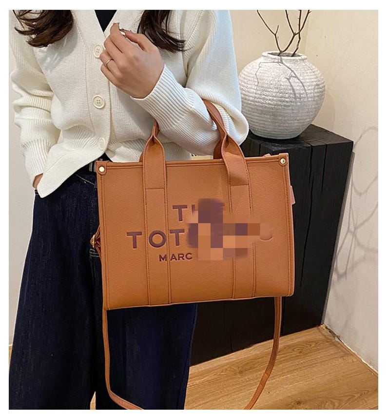 May include: A brown tote bag with top handles and a shoulder strap. The bag has the words "TOT" and "MARC" printed on the front. The bag is made of a smooth material and has gold-colored hardware.