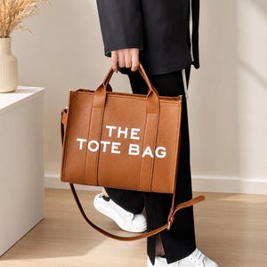 Pu&ograve; includere: Una borsa tote marrone con la scritta "THE TOTE BAG" in bianco. La borsa ha due manici e una tracolla. La borsa &egrave; tenuta da una persona che indossa pantaloni neri e scarpe da ginnastica bianche.