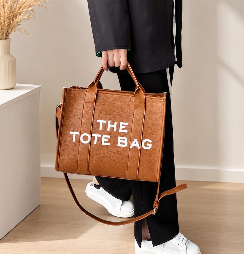 Pu&ograve; includere: Una borsa tote marrone con la scritta "THE TOTE BAG" in bianco. La borsa ha due manici e una tracolla. La borsa &egrave; tenuta da una persona che indossa pantaloni neri e scarpe da ginnastica bianche.
