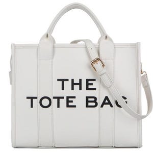 Pu&ograve; includere: Borsa tote bianca con la scritta "THE TOTE BAG" in nero. La borsa ha un manico superiore e una tracolla staccabile con fibbia color oro. La borsa &egrave; rettangolare e ha un design semplice e pulito.