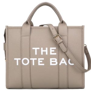 Pu&ograve; includere: Una borsa tote beige con la scritta "THE TOTE BAG" in lettere bianche. La borsa ha due manici e una tracolla staccabile con fibbia color oro. La borsa &egrave; realizzata in un materiale strutturato.