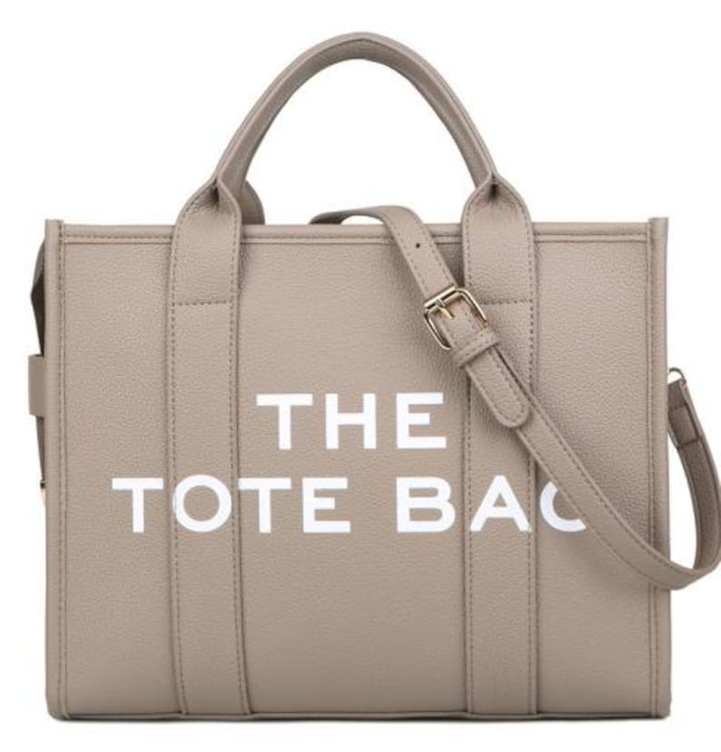 Pu&ograve; includere: Una borsa tote beige con la scritta "THE TOTE BAG" in lettere bianche. La borsa ha due manici e una tracolla staccabile con fibbia color oro. La borsa &egrave; realizzata in un materiale strutturato.