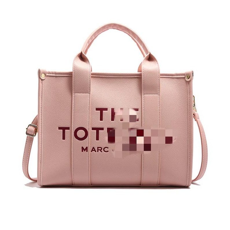 May include: A light pink tote bag with the words "THE TOTE" and "MARC" printed on the front. The bag has two handles and a detachable shoulder strap. The hardware is gold-colored.