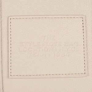 Pu&ograve; includere: Primo piano di una borsa in pelle color crema con una toppa rettangolare cucita sul davanti. La toppa presenta un testo in rilievo e la borsa ha dettagli di cucitura verticali.