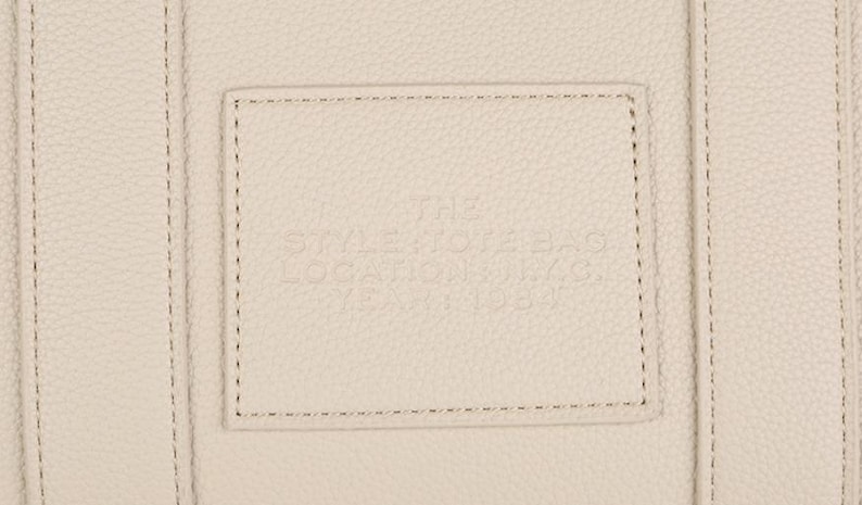 Pu&ograve; includere: Primo piano di una borsa in pelle color crema con una toppa rettangolare cucita sul davanti. La toppa presenta un testo in rilievo e la borsa ha dettagli di cucitura verticali.