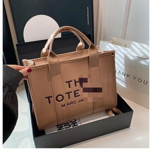 May include: A tan tote bag with the words "THE TOTE MARC JA" printed on the front. The bag has two handles and a small strap. It is sitting in a black box.