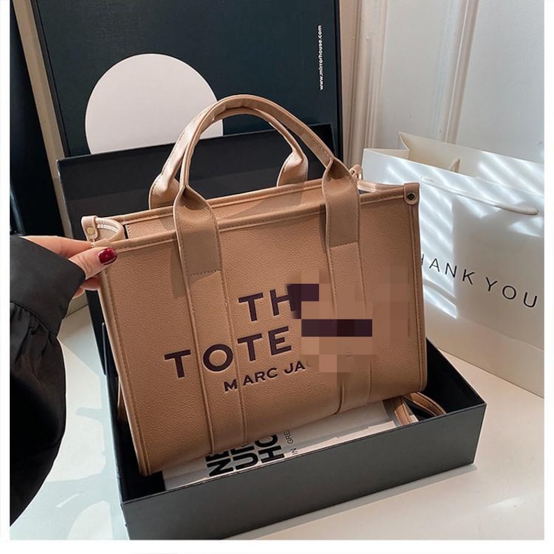 May include: A tan tote bag with the words "THE TOTE MARC JA" printed on the front. The bag has two handles and a small strap. It is sitting in a black box.