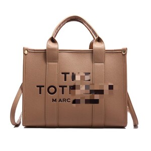 May include: A tan tote bag with the words "THE TOTE MARC" printed on the front. The bag has two top handles and a detachable shoulder strap. The bag is made of a smooth material and has a rectangular shape.