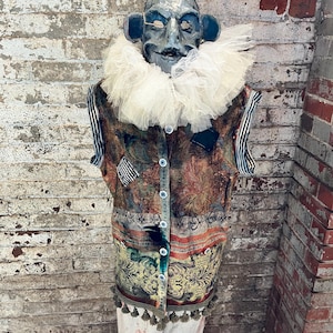 May include: A mannequin head with a grey, textured face and pointed ears wears a large, white, ruffled tulle collar. The torso is dressed in a sleeveless, multi-colored tapestry vest featuring ornate patterns, button closures, and a bottom fringe.