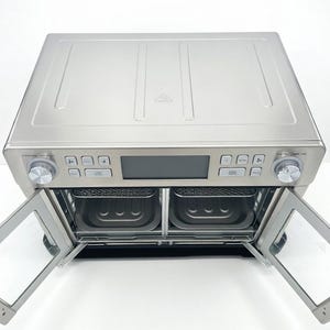 May include: A stainless steel air fryer oven with two pull-down doors. The appliance has a digital display and control knobs. The interior reveals two cooking baskets. The doors are clear, allowing a view of the interior.
