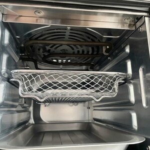 May include: Interior view of a stainless steel air fryer oven. The image shows the inside components, including a wire mesh basket, a heating element, and a drip tray. The oven is empty and ready for use.