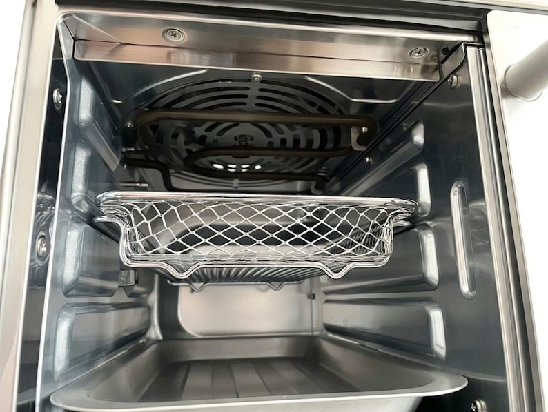 May include: Interior view of a stainless steel air fryer oven. The image shows the inside components, including a wire mesh basket, a heating element, and a drip tray. The oven is empty and ready for use.