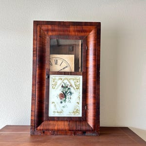 Puede incluir: Un reloj de pared rectangular de madera con un acabado de veta de madera marrón oscuro. La sección superior presenta un panel de vidrio que revela una esfera parcial con números romanos. La sección inferior contiene un panel de vidrio blanco.