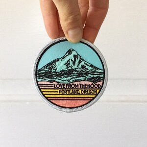 May include: Round patch with a mountain design and the text "LOVE FROM THE HOOD PORTLAND, OREGON." The patch features a mountain in black, a blue sky, and a colorful sunset design with a white border.