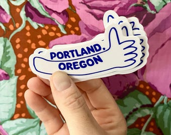 May include: White sticker with a blue outline of a hand giving a thumbs up. The sticker says "PORTLAND, OREGON" in blue text.