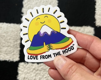 May include: A white sticker with a yellow sun smiling and hugging a purple and blue mountain. The text "LOVE FROM THE HOOD™" is written below the sun and mountain.