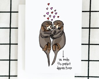 May include: A greeting card with a hand-drawn illustration of two otters hugging. Pink hearts float above them. The text below reads "we make the perfect dream team".