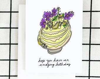 May include: A white card with a hand-drawn illustration of a yellow cake with purple flowers and the text "hope you have an amazing birthday".