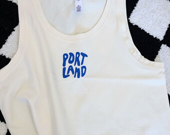 May include: A white tank top with a blue "PORTLAND" graphic on the front. The tank top is cropped and has a scoop neck.
