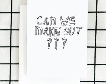 May include: A white greeting card with the text "CAN WE MAKE OUT ???" in black, handwritten-style lettering. The card is paired with a blank white envelope, set against a black and white checkered background.