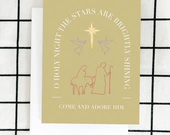 May include: A beige Christmas card with the text "O Holy Night The Stars Are Brightly Shining Come And Adore Him". The card features a star with two angels and a depiction of Mary, Joseph, and the donkey.