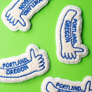 May include: Four white felt patches with blue embroidery. Each patch features the text "Portland, Oregon" and a stylized hand giving a thumbs up.