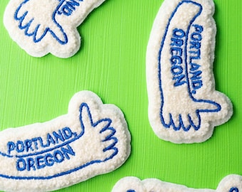 May include: Four white felt patches with blue embroidery. Each patch features the text "Portland, Oregon" and a stylized hand giving a thumbs up.