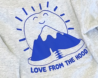 May include: A gray sweatshirt with a blue graphic of a smiling sun hugging a mountain. The text "LOVE FROM THE HOOD" is printed below the graphic.