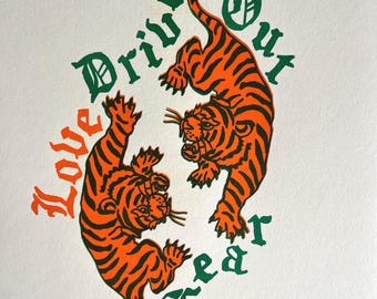 May include: Two orange and black tigers facing each other with green text that reads "Love Drives Out Fear".