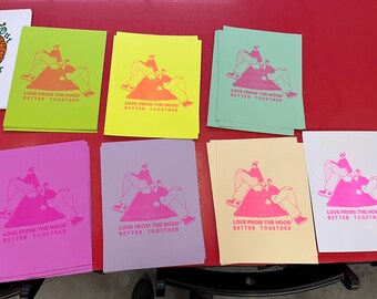 May include: A collection of colorful art prints featuring a graphic illustration of two figures lounging on a mountain. Each print has the text "LOVE FROM THE HOOD BETTER TOGETHER" in a matching color. The prints are in various colors, including green, yellow, pink, and purple.