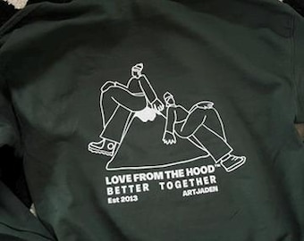 May include: A dark green hoodie with a white graphic of two figures sitting on a mountain. The text "LOVE FROM THE HOOD" BETTER TOGETHER" is printed below the graphic. The text "Est 2013" and "ARTJADEN" are printed below the main text.