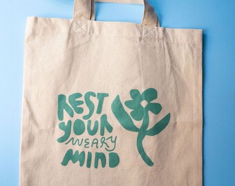 May include: A beige canvas tote bag with green text and a green flower design. The text reads "Rest your weary mind".