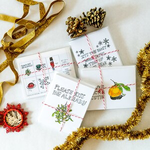 May include: A set of six Christmas cards featuring hand-drawn illustrations. The cards are tied together with red and white twine. The cards feature illustrations of mistletoe, a nutcracker, a Christmas pudding, and a tangerine. The text on the cards includes "Please Kiss Me Already", "It's the Most Wonderful Time of Year", and "Can You Be My Cutie".