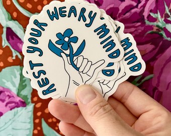 May include: A stack of white stickers with blue lettering and a blue flower graphic. The stickers say "Rest Your Weary Mind".
