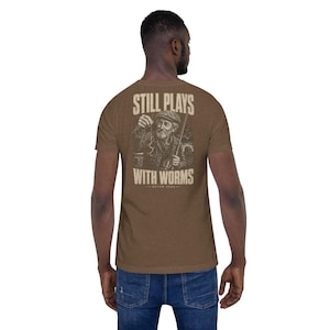 Pode incluir: Camiseta marrom com um gráfico de um homem mais velho pescando, segurando uma minhoca. O texto "STILL PLAYS WITH WORMS" está acima e abaixo da ilustração. A camiseta é feita de um material macio.