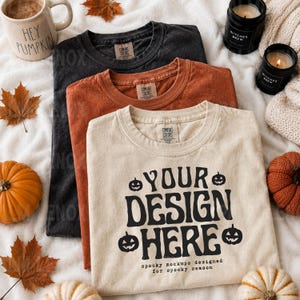 Op de afbeelding: Drie T-shirts met ronde hals in zwart, roestoranje en crème. Het crèmekleurige shirt heeft zwarte tekst "YOUR DESIGN HERE" omringd door vier pompoenpictogrammen, met daaronder de kleinere tekst "spooky mockups designed for spooky season."
